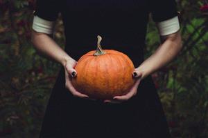 Woman in a black dress with black painted fingernails holding a pumpkin.